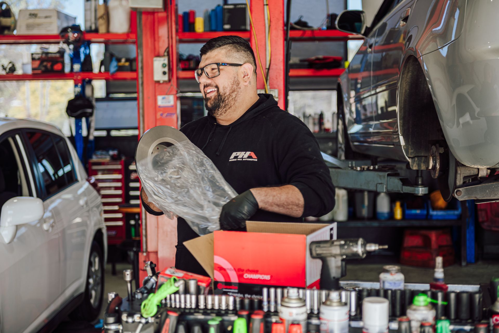 Mechanic in a garage holds part, smiling. Car parts on table, vehicle on lift.