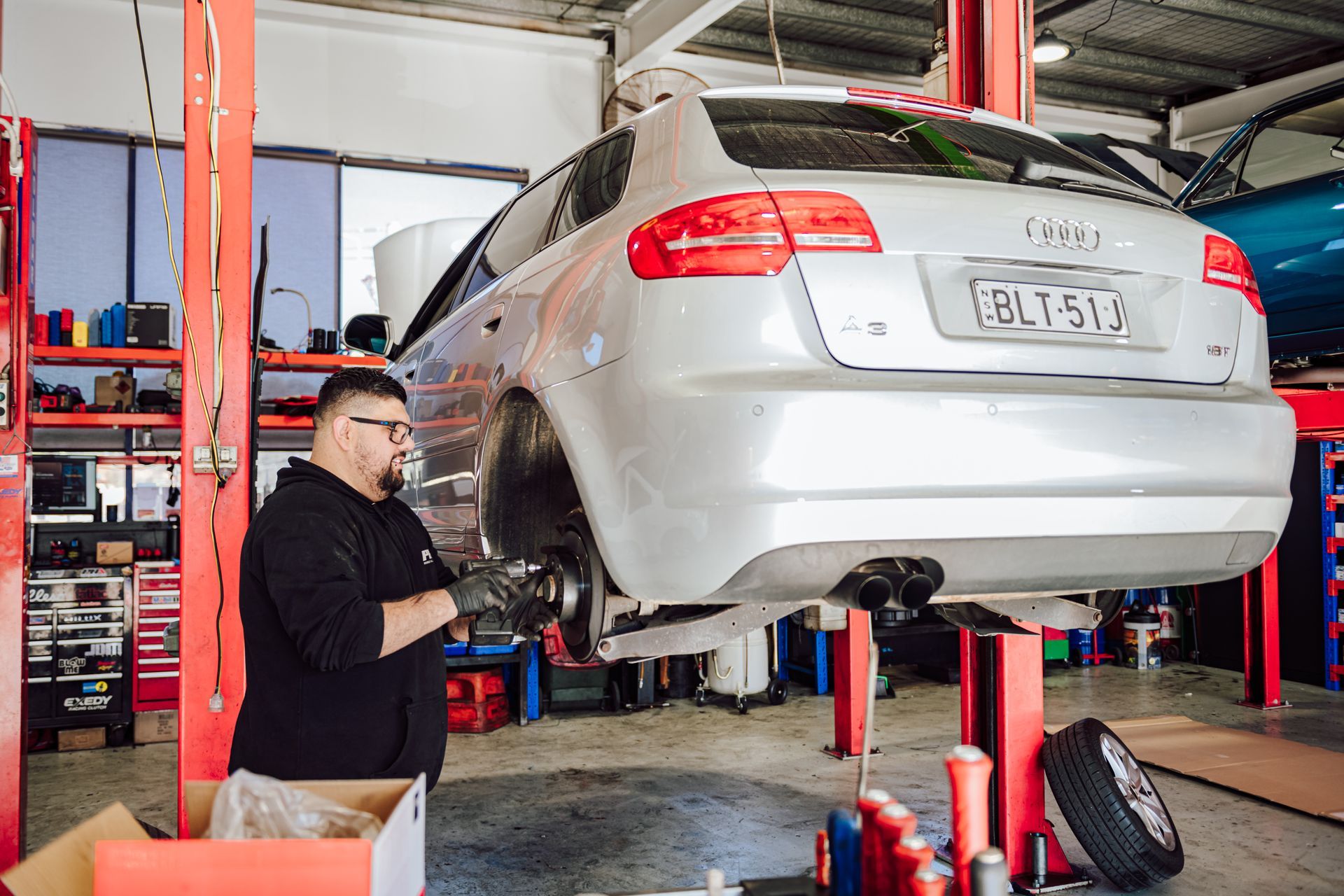 Mechanic working on a silver Audi in a repair shop; the car is lifted on a red lift.