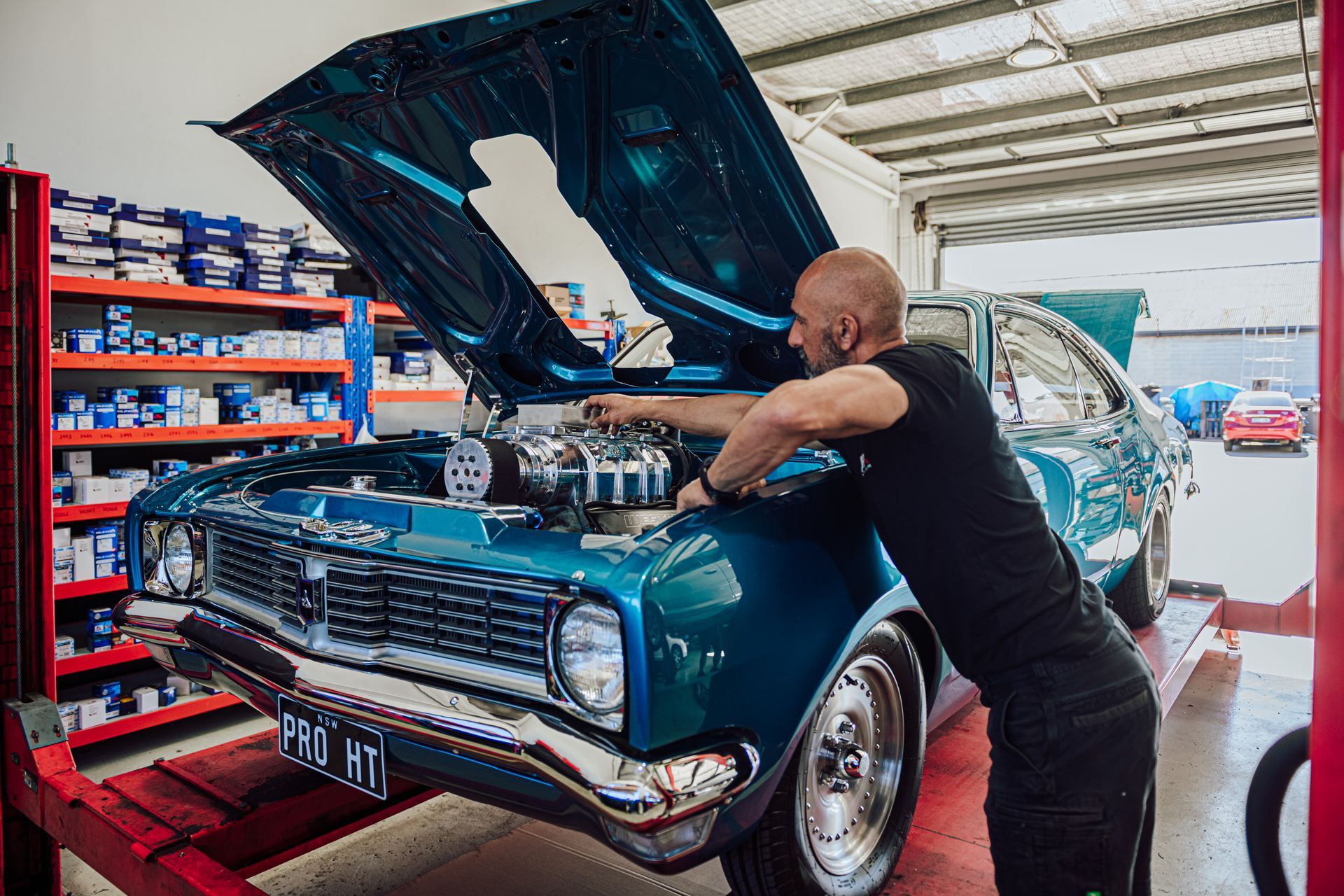 Mechanic working on a teal classic car with the hood up in a garage.