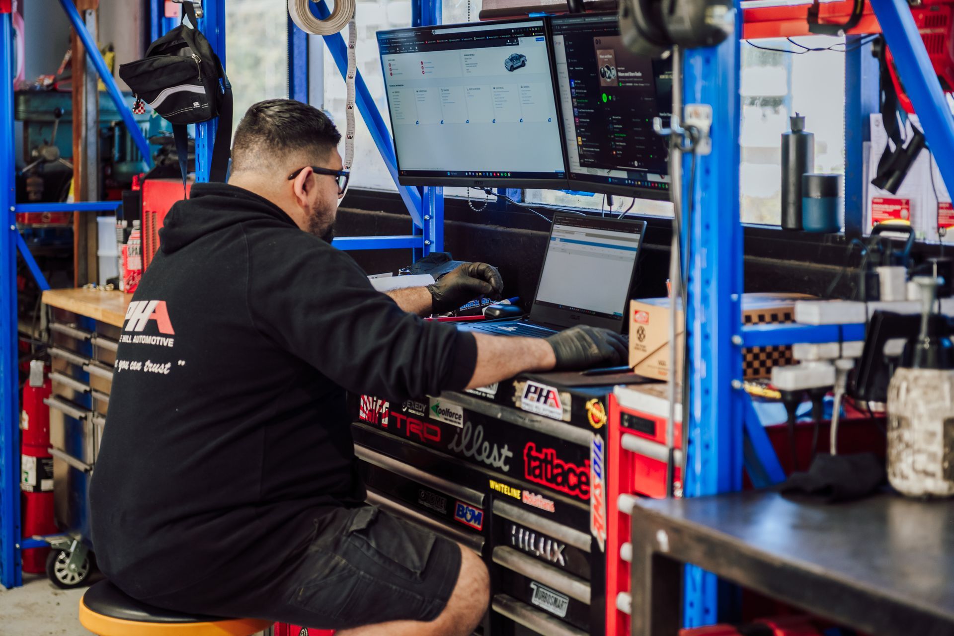 Mechanic at computer, working on a laptop with dual monitors, in a garage setting.
