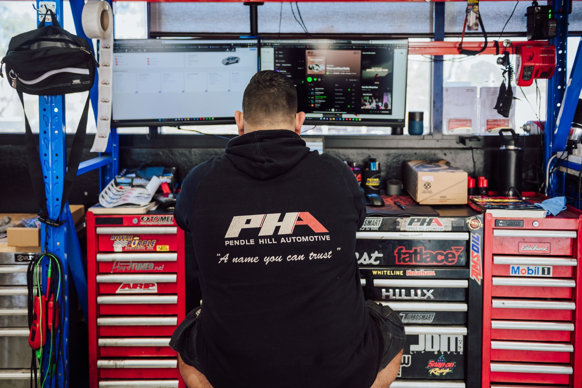 Man in black hoodie working at a workbench with computer monitors. Workshop setting with red tool chests.