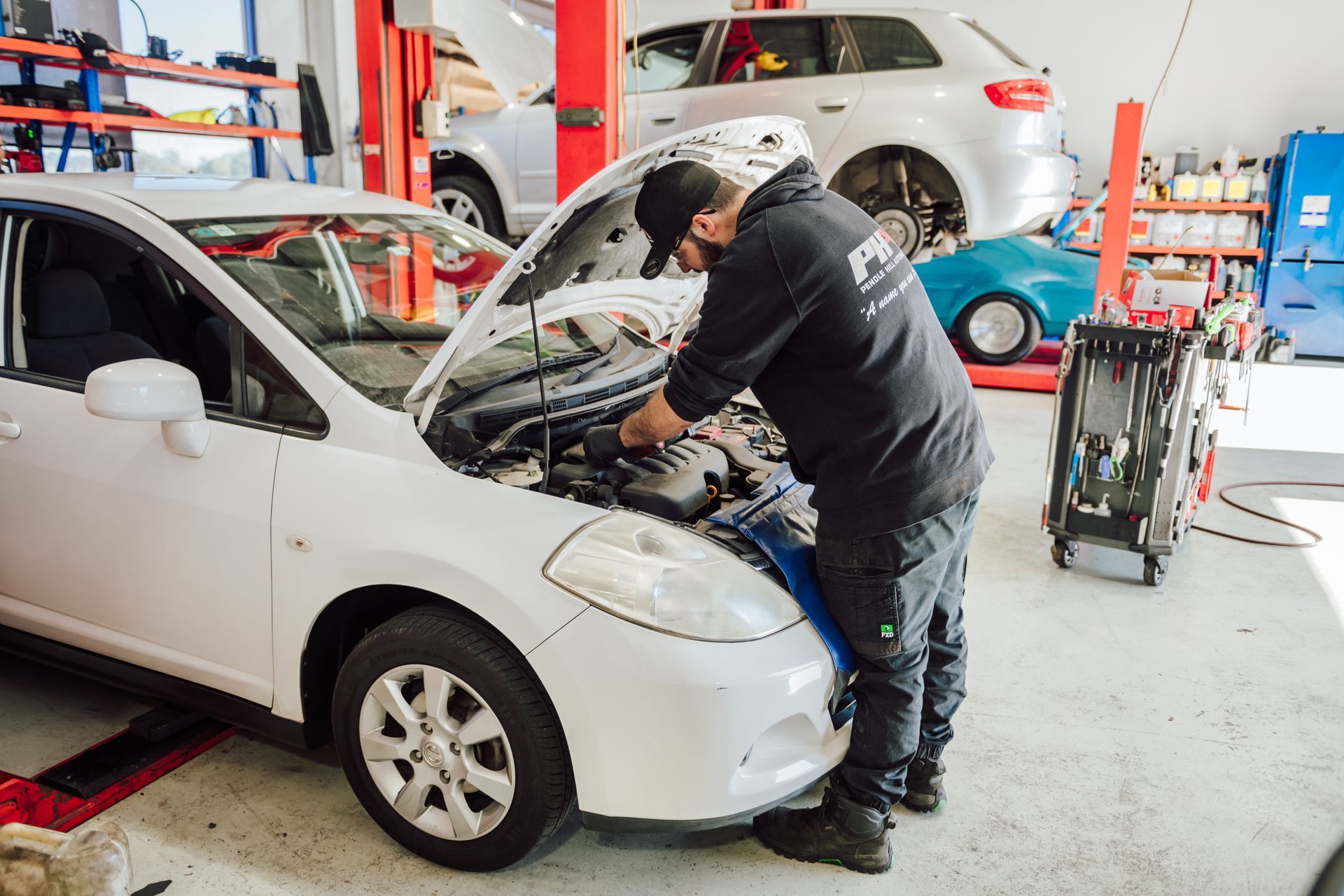 Mechanic working on a white car with the hood open inside a car repair shop.
