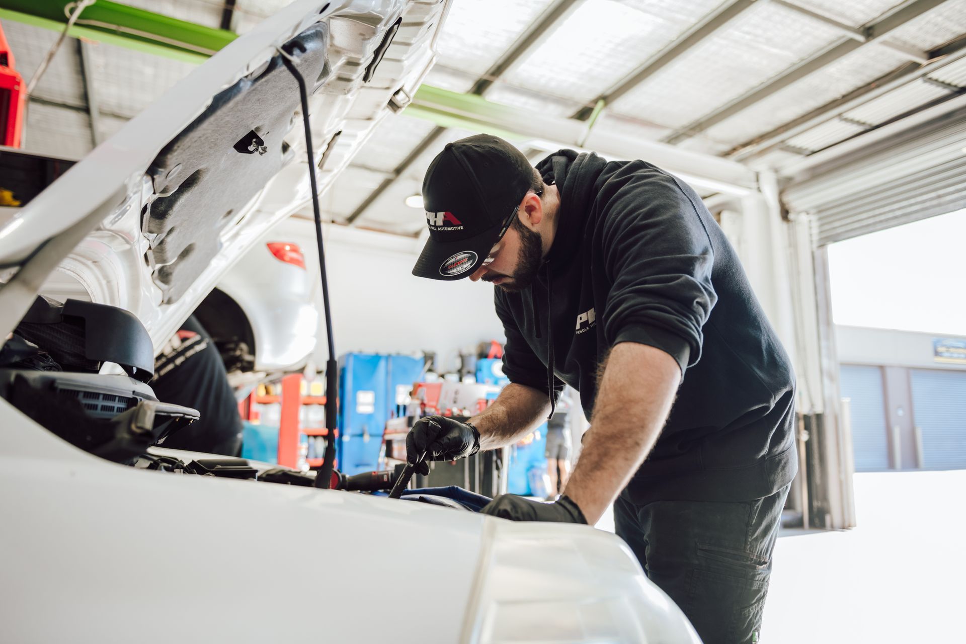Mechanic works on a car engine in a garage; wearing black hat, gloves, and hoodie.