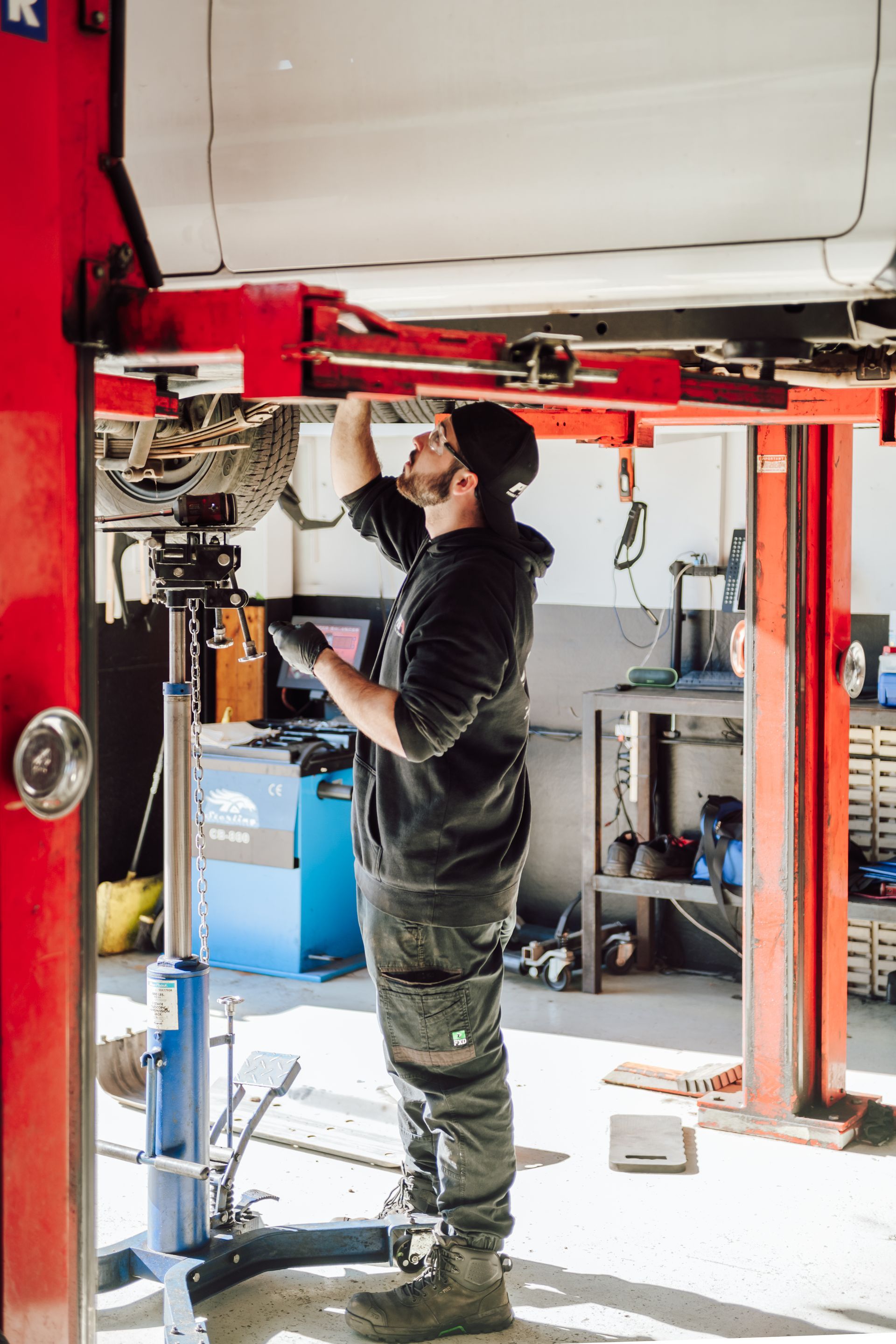 Mechanic working under a car raised on a lift in a garage. He is wearing a black hoodie and looking up.