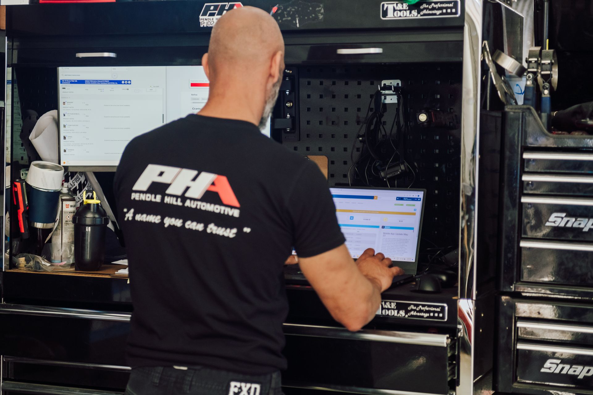 Man at a computer in a garage; he is wearing a black shirt, working on a laptop and a monitor.