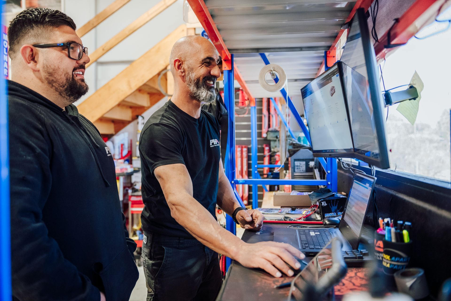 Two men in a shop, looking at a laptop and monitor, smiling. Blue and red shelving.
