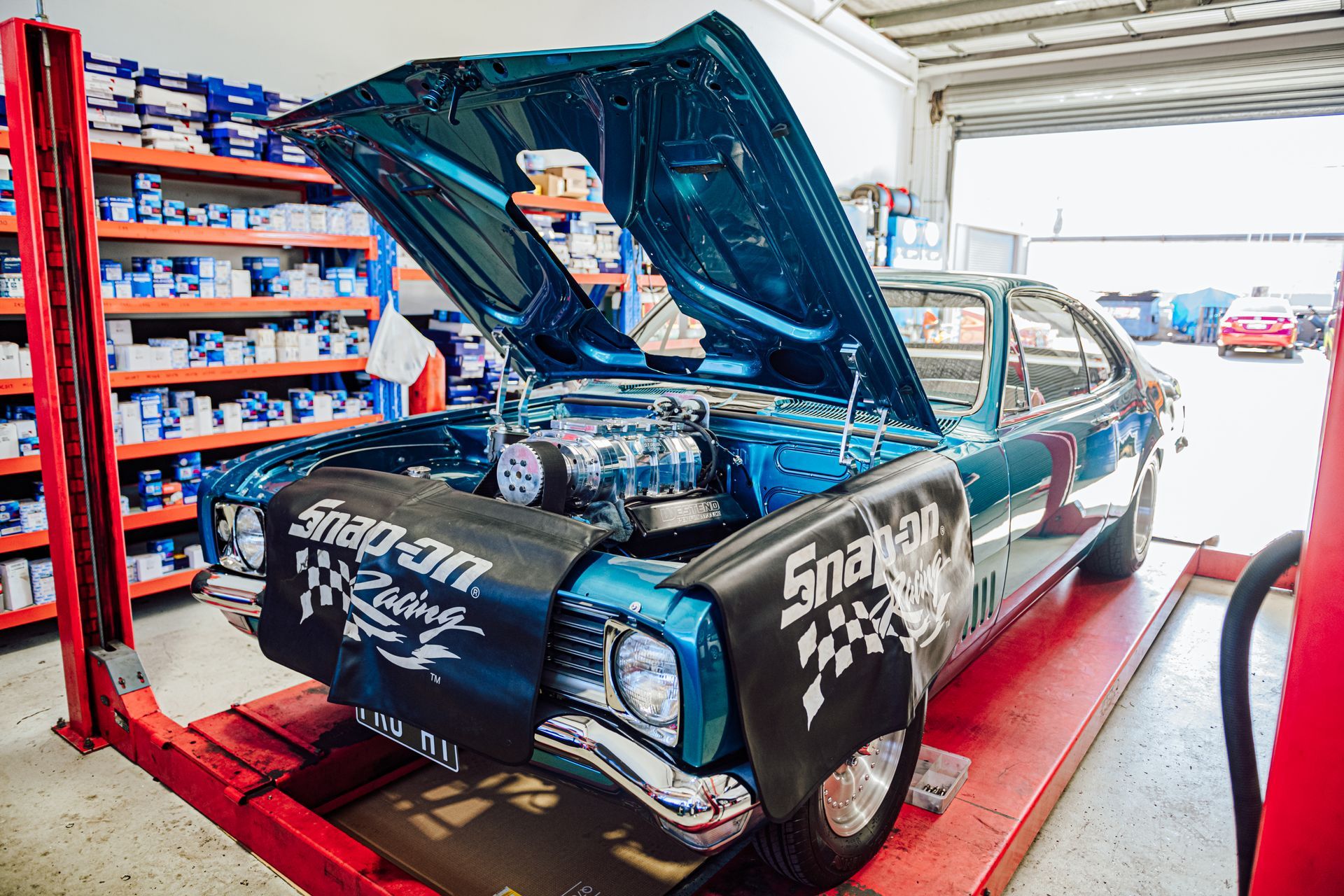 Blue classic car on a lift, hood open in a repair shop, Snap-on Racing fender covers.