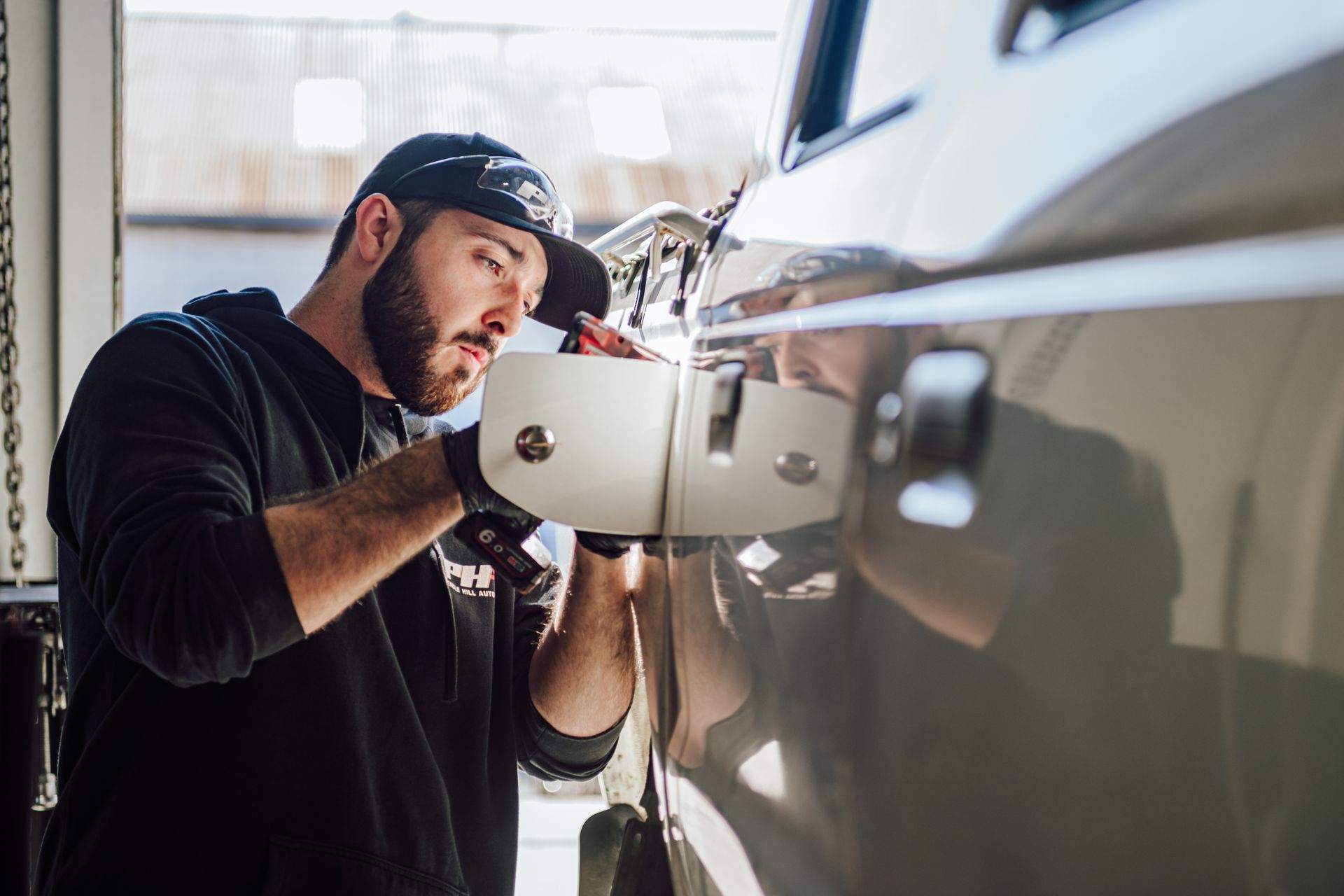 Mechanic installing a white side mirror on a gray vehicle in a workshop.
