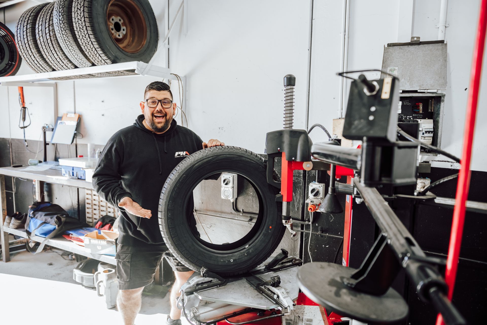 Man in garage holding a tire, smiling. Tire machine nearby. Tires on shelf, work bench.