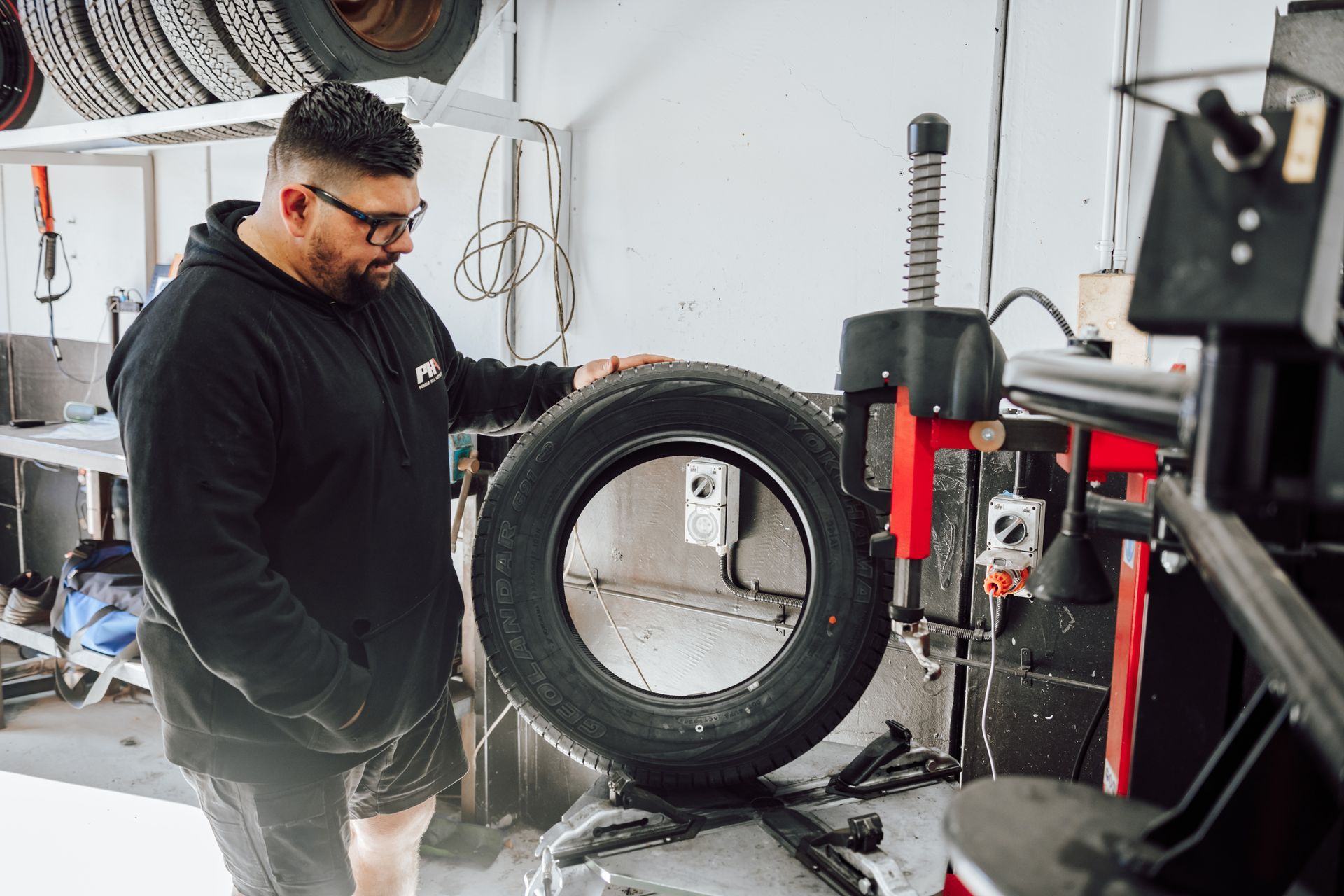 Man in a garage examining a tire on a machine, other tires on the wall.