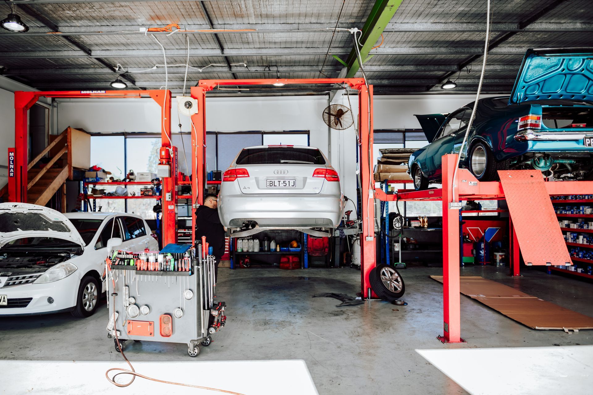 A mechanic working on a car lifted in a well-lit auto repair shop; other cars, tools visible.