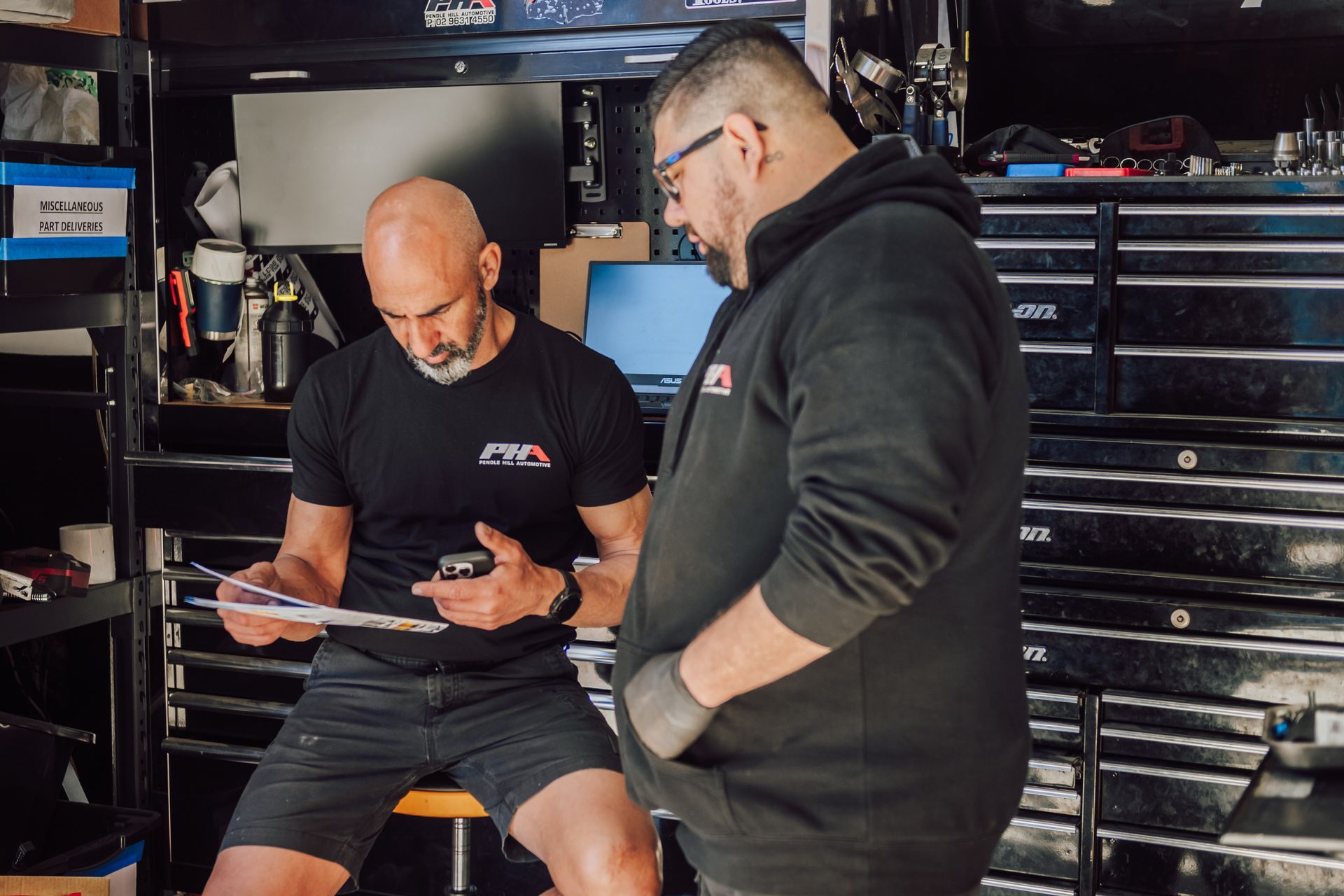 Two men in a garage looking at paperwork. One bald, in black, the other in a hoodie, looking down.