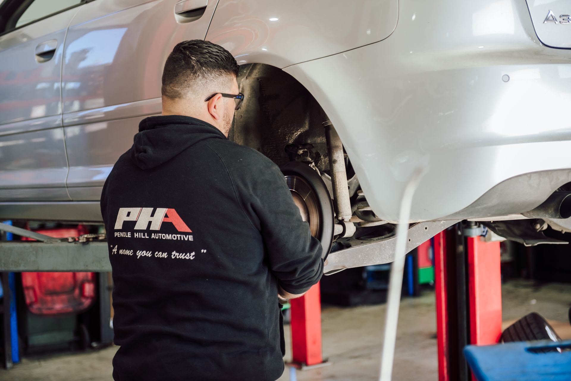 Mechanic working on car's rear wheel, wearing a black hoodie, in a shop.