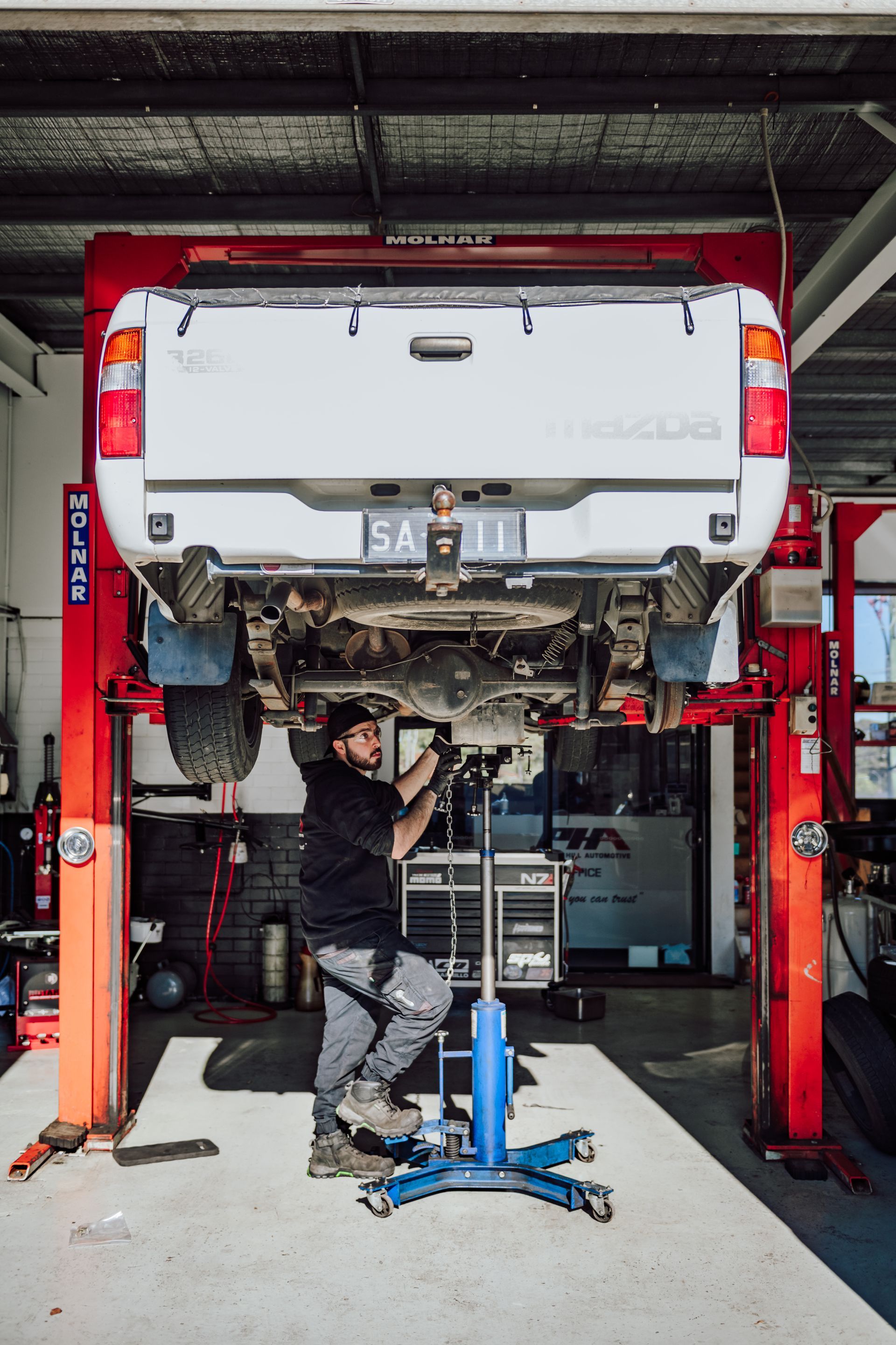 Mechanic working on white pickup truck hoisted in a garage. Red lift, concrete floor, man wearing work clothes.