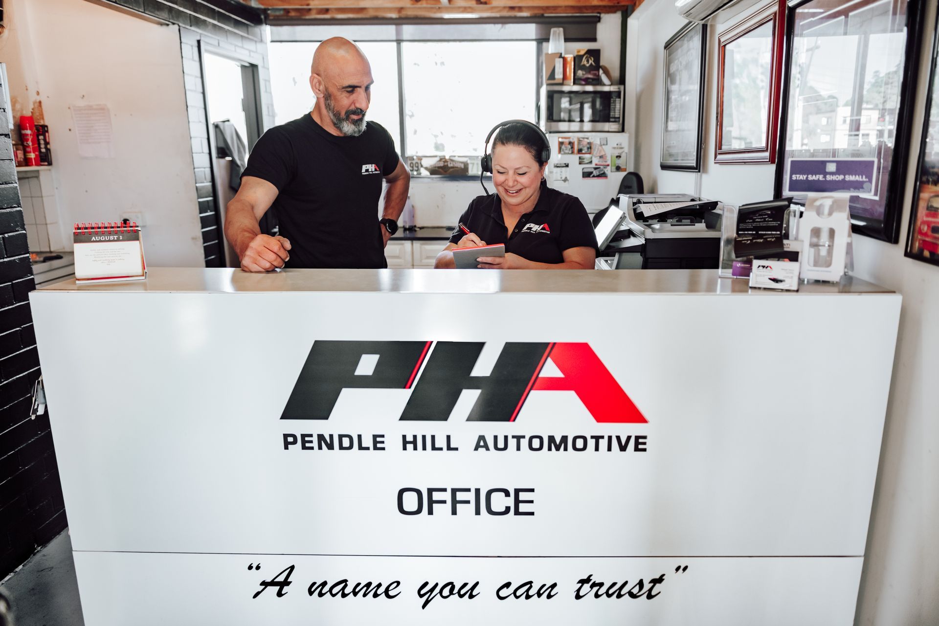Two people at the PHA Pendle Hill Automotive office. A man and woman stand behind a white desk.