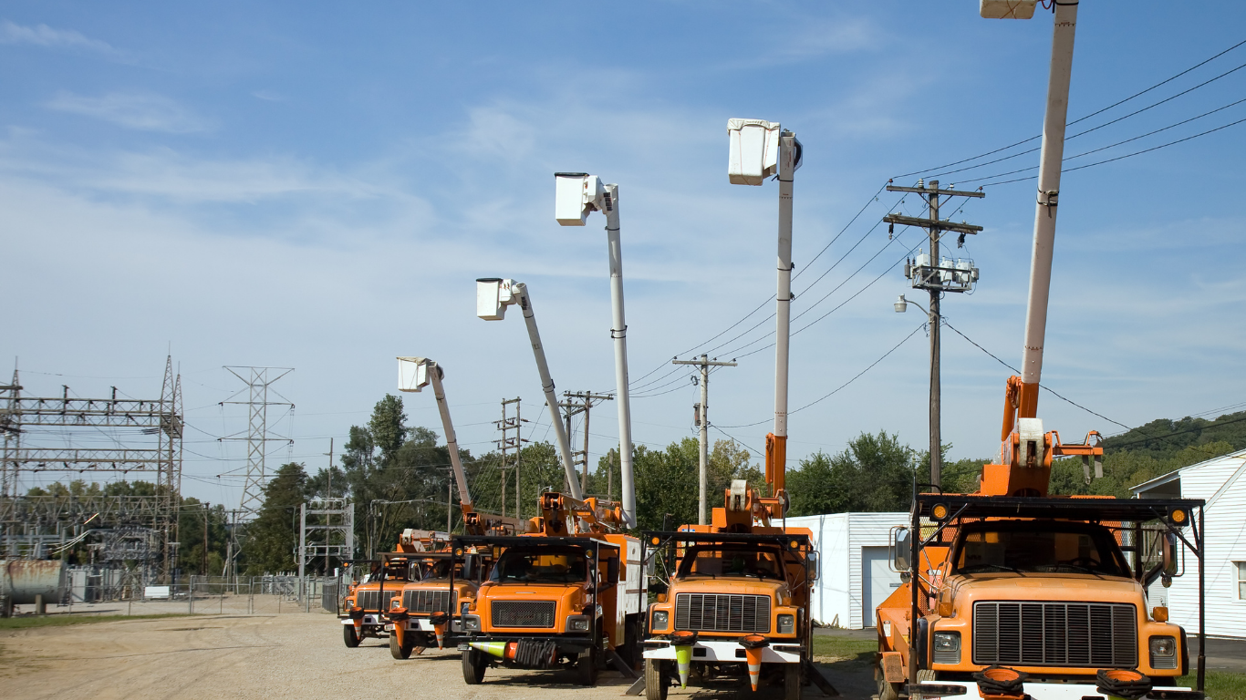 Orange utility trucks with raised buckets near power lines and a substation on a sunny day.