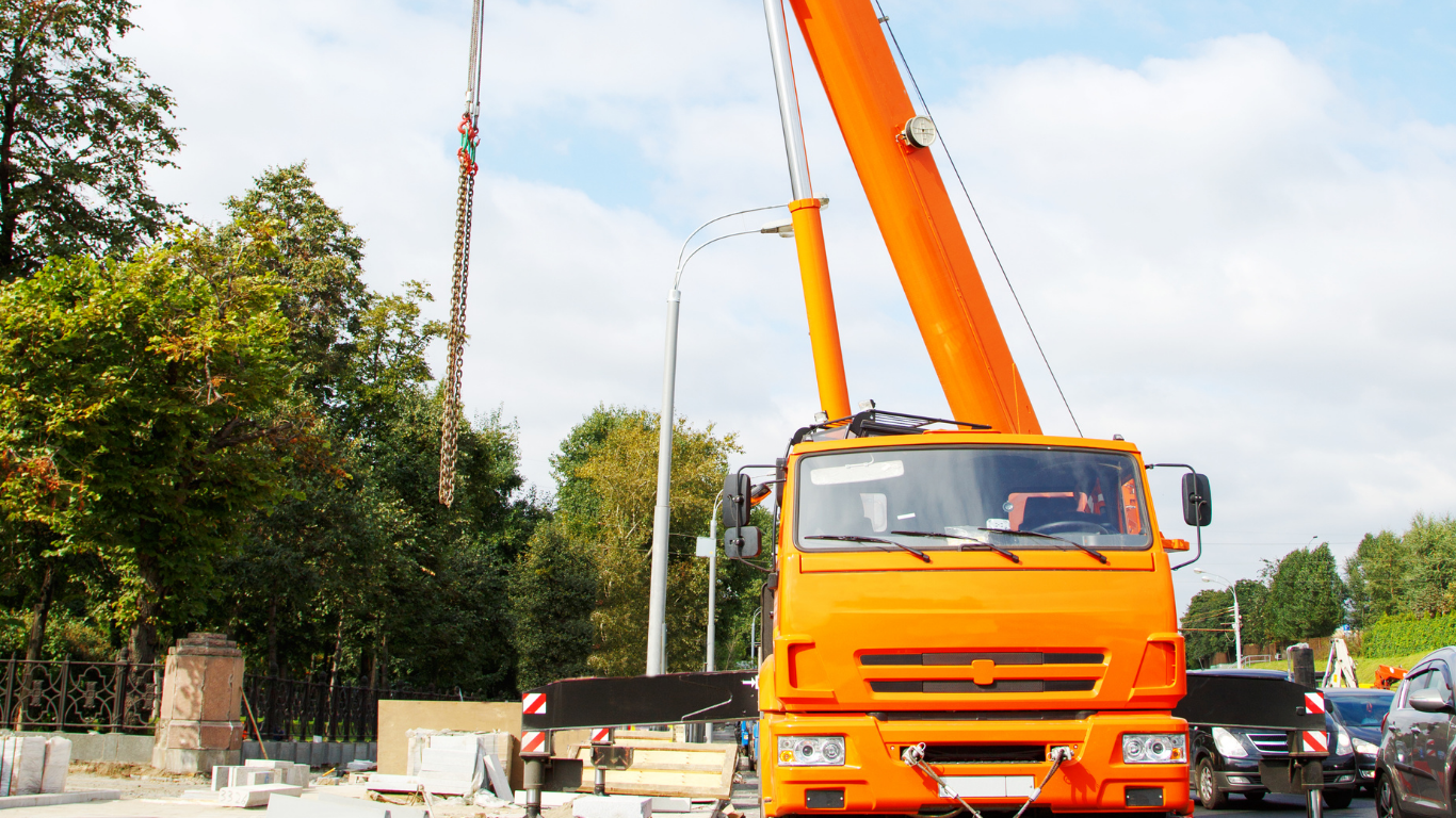 Orange crane lifting a long structure; trees and cars in the background.