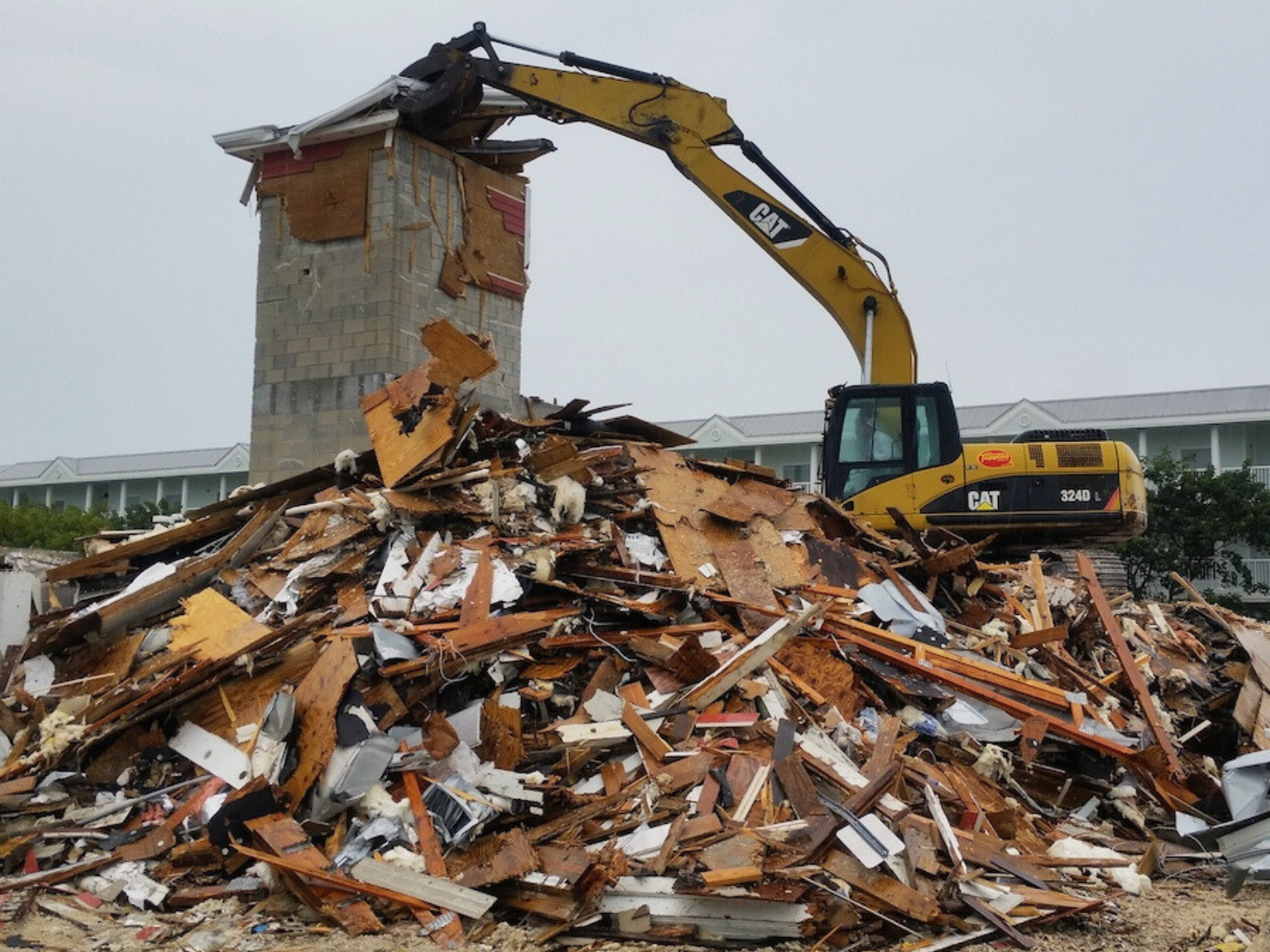 An excavator is actively tearing down the remaining portion of a concrete block tower.