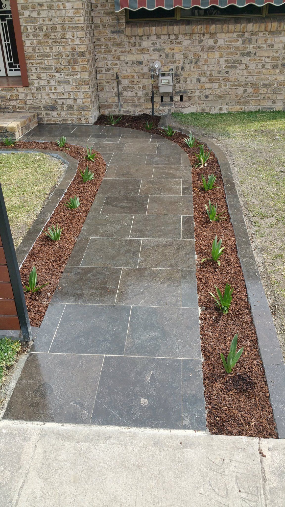 A walkway leading to a house with a brick wall and a few plants on it.