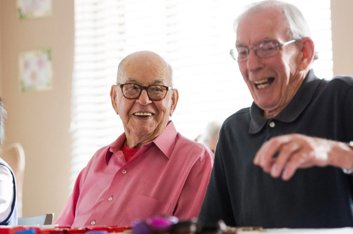 Two older men laughing indoors, one in a pink shirt, the other in a black polo shirt.