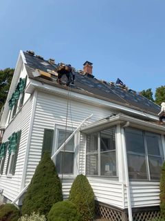 Workers are installing new shingles on the roof of a white two-story house on a sunny day.