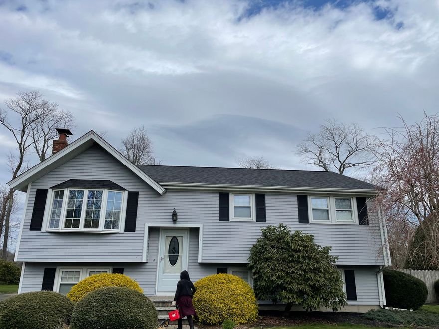 A light-gray split-level house with dark shutters, a front door, and three rounded shrubs, under a cloudy, overcast sky.