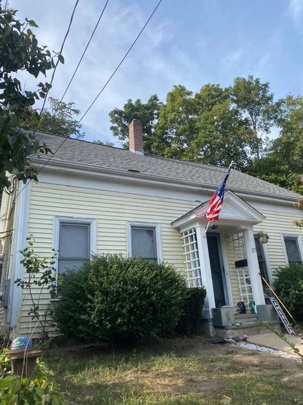 A one-story yellow house with a gabled roof, a brick chimney, and an American flag hanging by the front door.