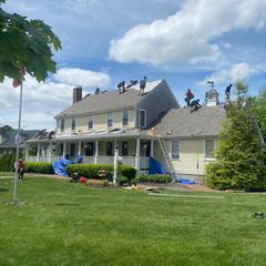 A crew of construction workers repairing the roof of a large, two-story yellow house under a bright, partly cloudy sky.