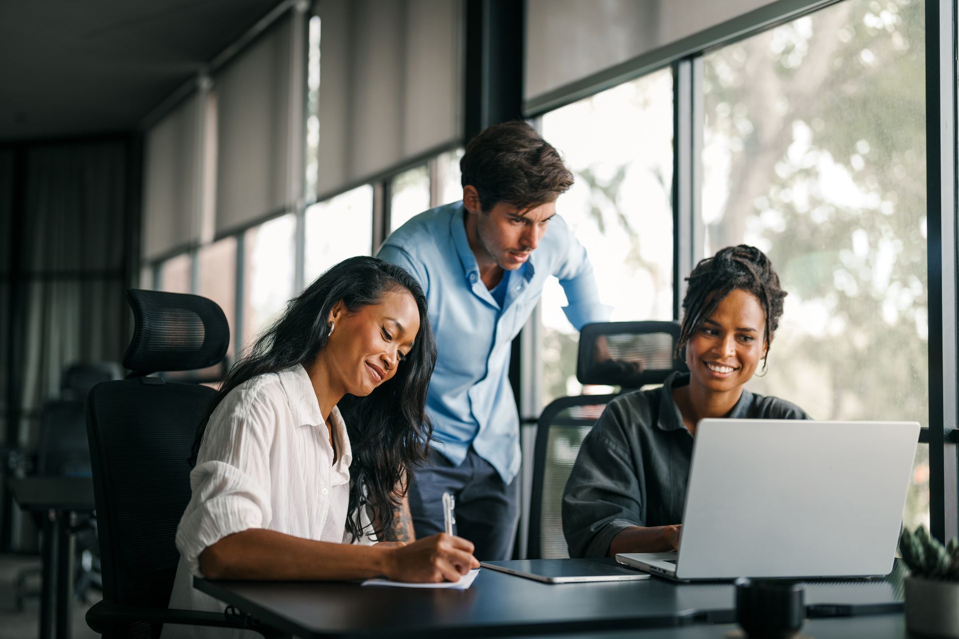Three people collaborating around a laptop in an office. One writes on paper, one types, and another looks on.