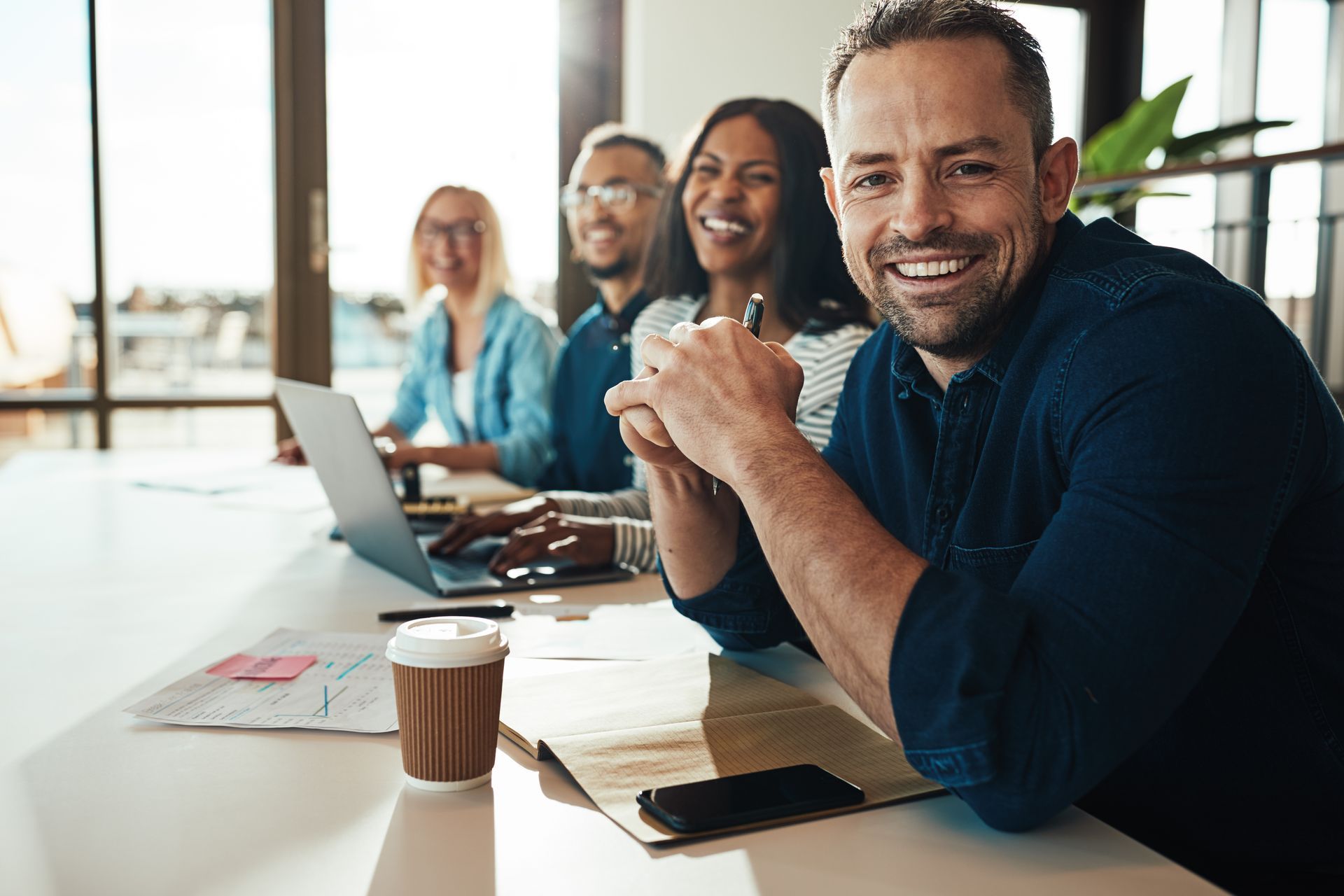 Office meeting: smiling man in blue shirt at a table with diverse colleagues, laptop, coffee, and papers.