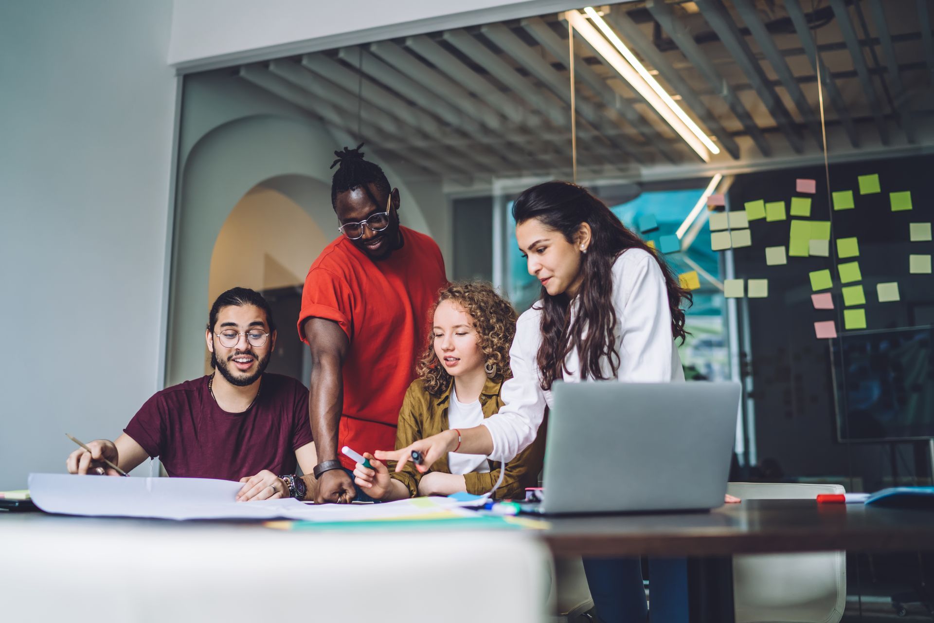 Four people collaborate around a table with papers and a laptop, pointing and discussing in an office setting.