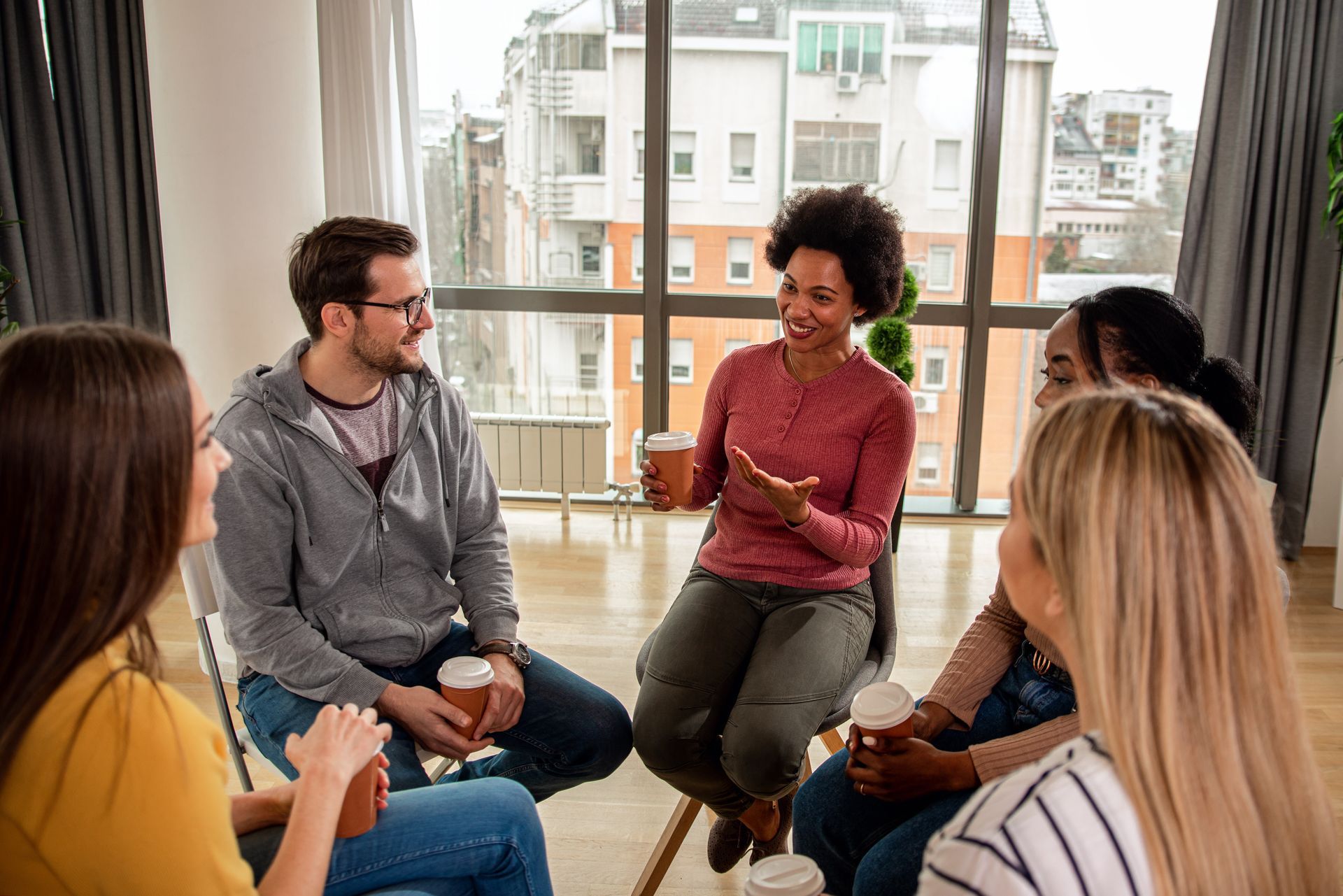 Group of people in a circle, conversing. A woman is speaking with animated gestures, all holding coffee cups. Bright, airy room.