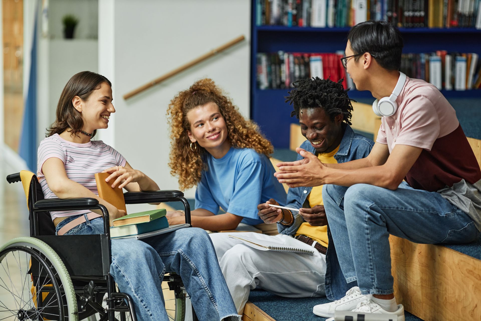 Four students chatting in a library setting, one using a wheelchair. They are smiling, with books and notes.