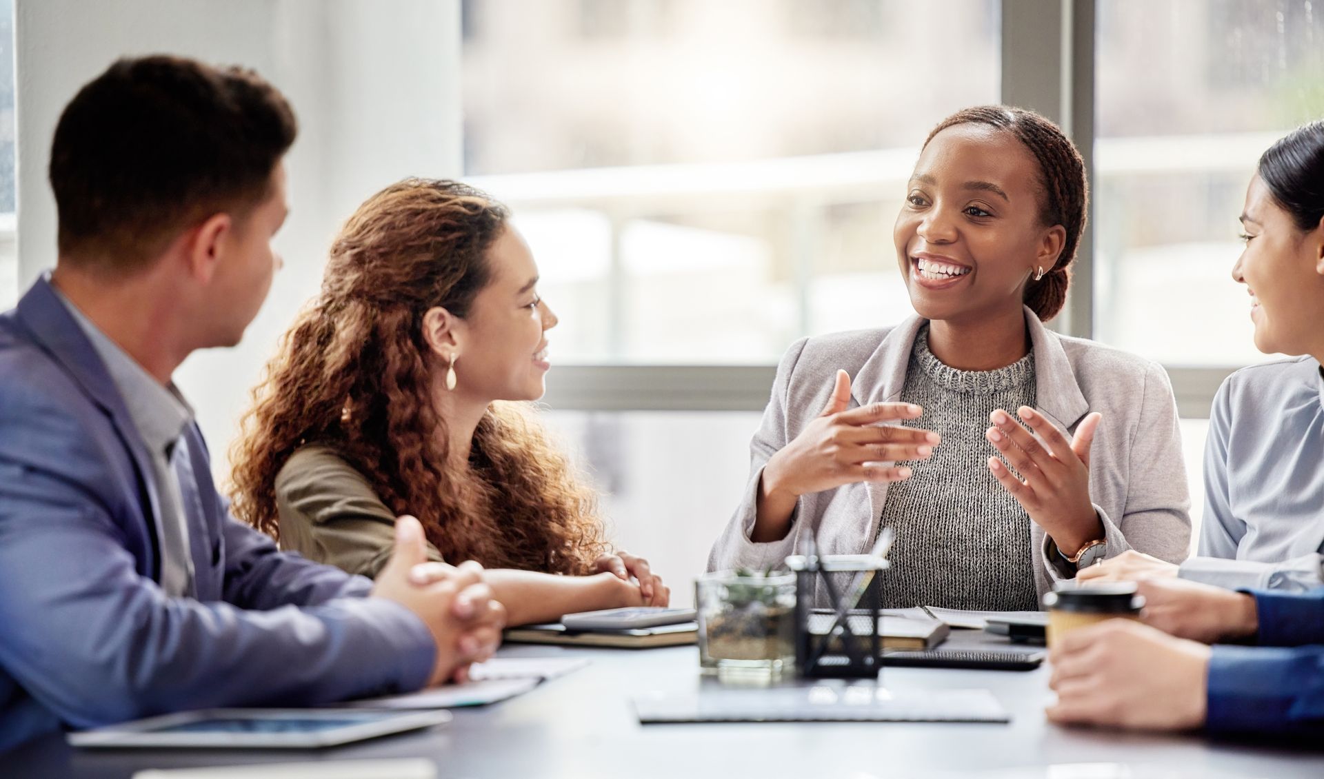 Business meeting around a table; a woman speaks animatedly, gesturing with hands; others listen.