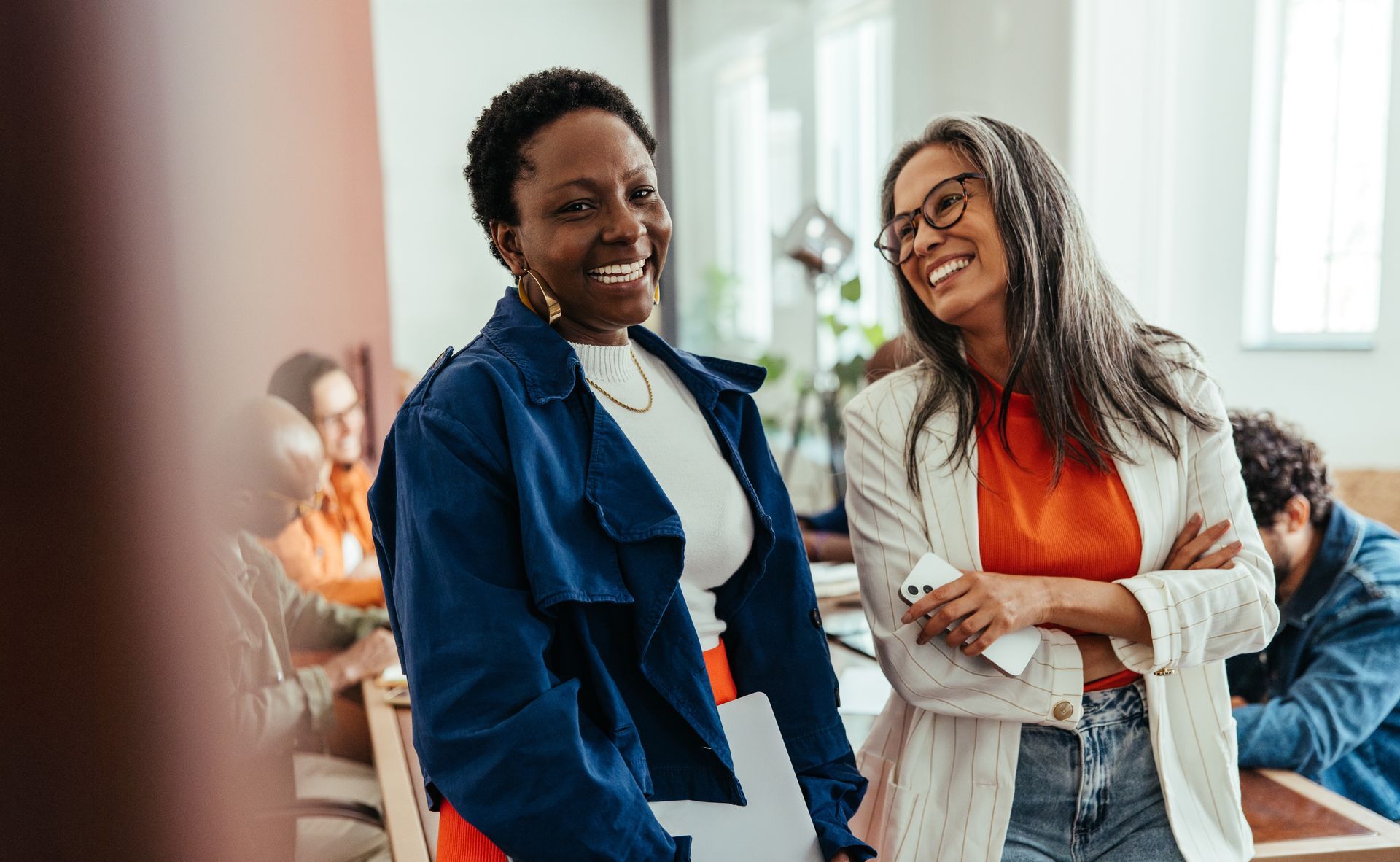 Two women smiling, chatting in an office. One in a blue jacket, one in a white blazer, others in background.