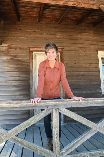 Woman sitting cross-legged in a field of dry grass, wearing a rust-colored top, jeans, and tan boots.