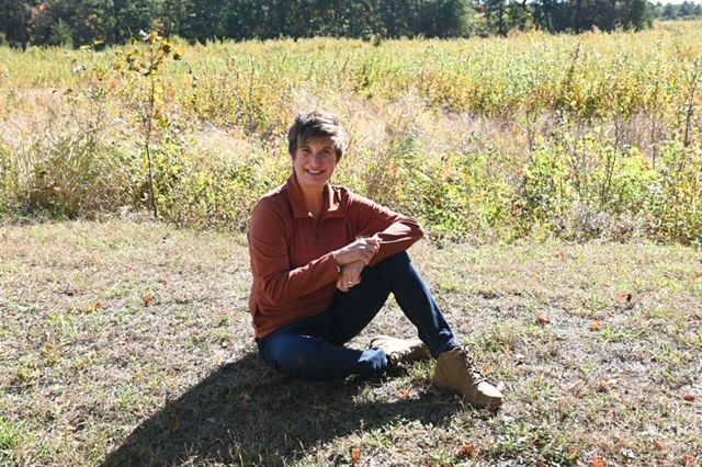 Woman in rust-colored sweater stands on porch of wooden cabin, leaning on railing, smiling.