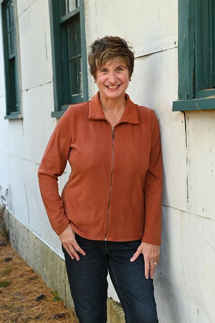Woman in rust-colored jacket and jeans, leaning against a white building, smiling.