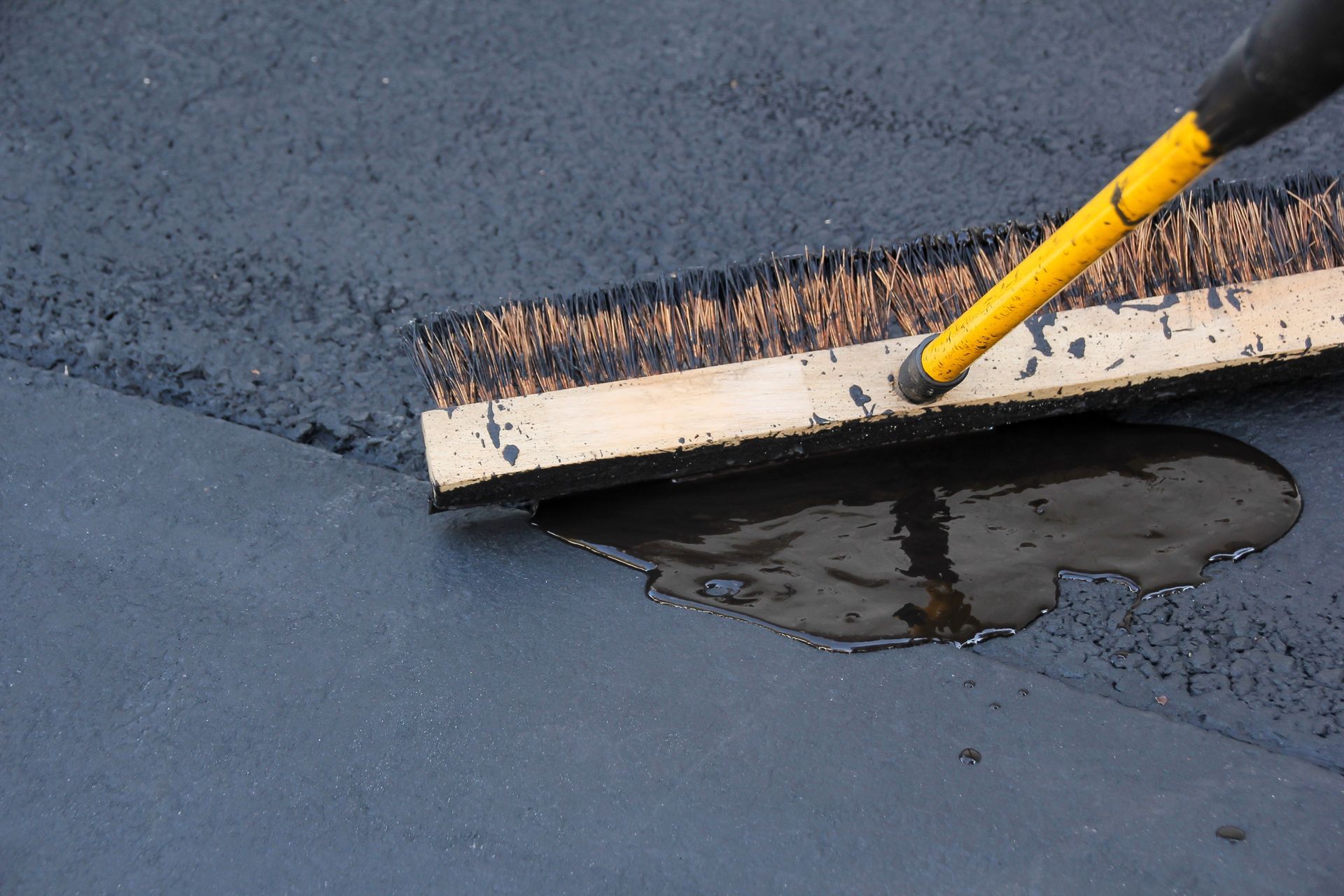 Person using a push broom to spread black asphalt. Yellow handle, brush is in contact with the asphalt surface.