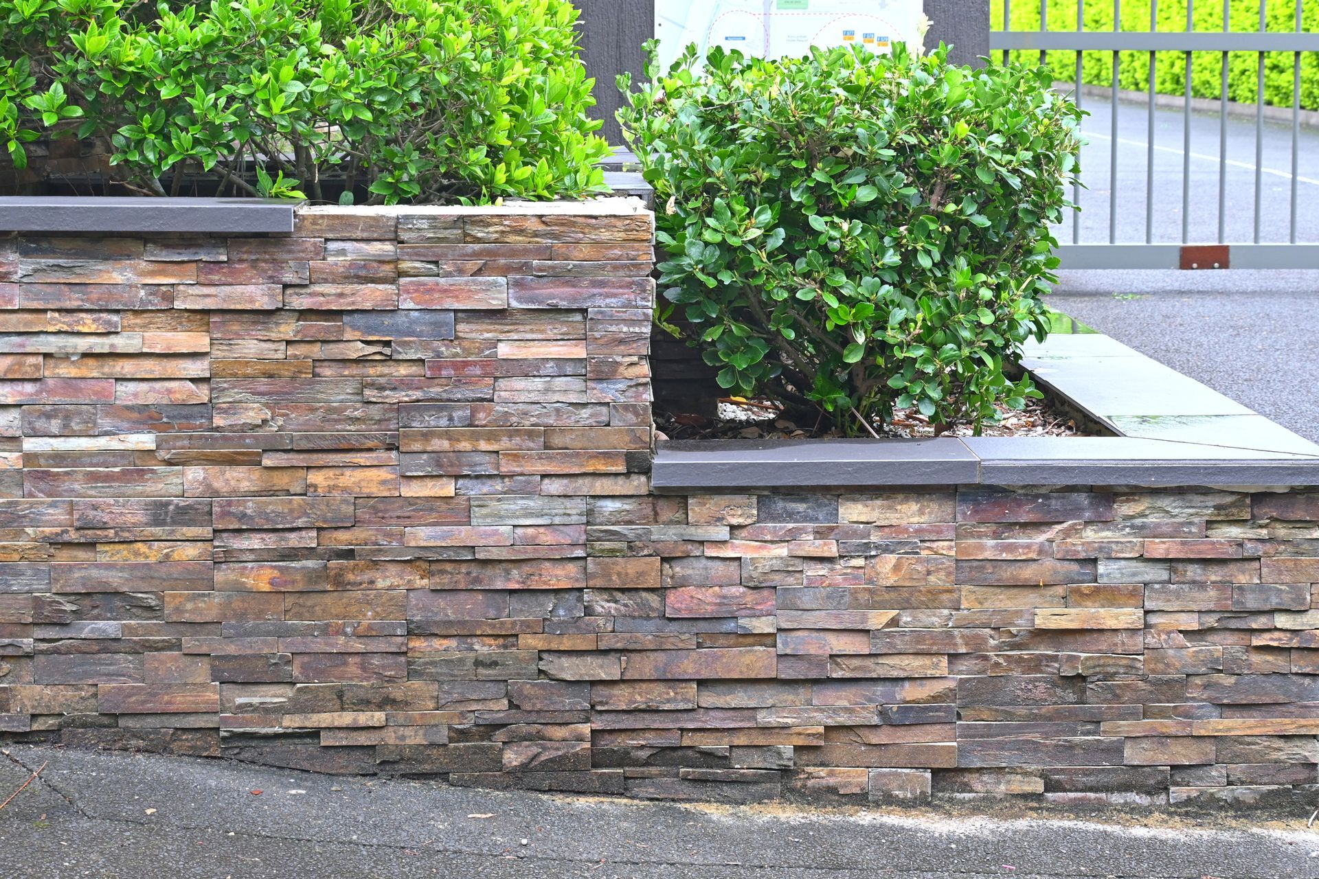 Stone veneer retaining wall with green bushes on a paved surface near a metal gate.