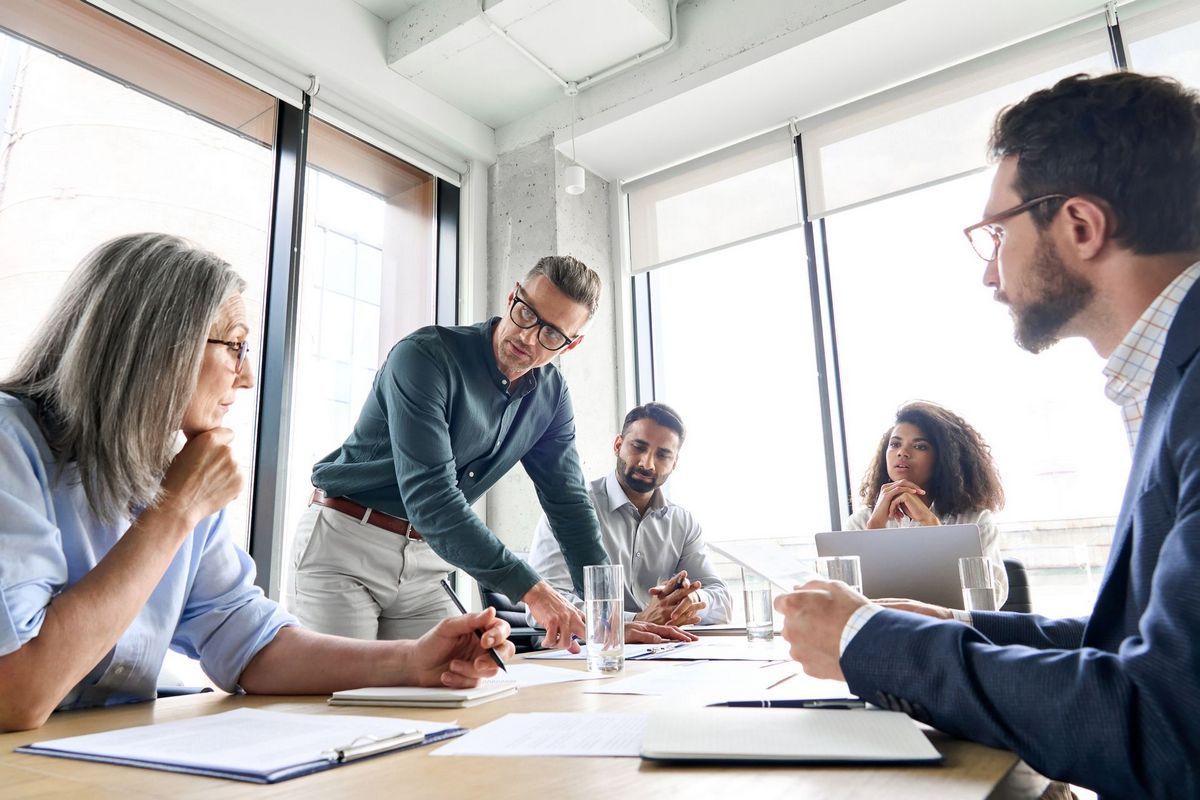 People in a bright office meeting around a table, discussing papers and laptops.
