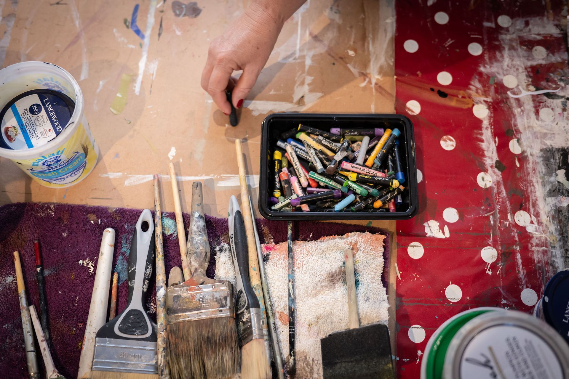 Hand holding charcoal, surrounded by art supplies on a paint-splattered table: brushes, crayons, and paint cans.