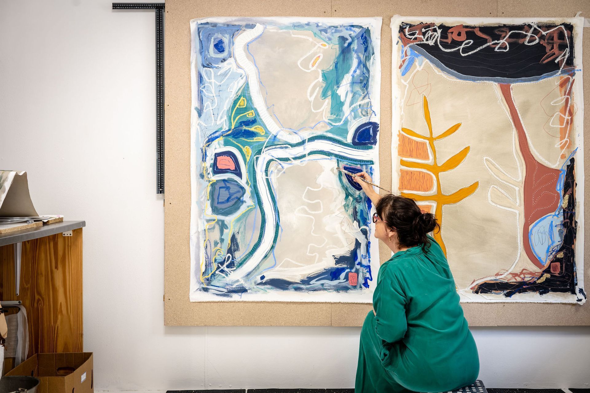 An artist in a green shirt paints on a large canvas featuring blue, white, and earthy abstract shapes in a studio.
