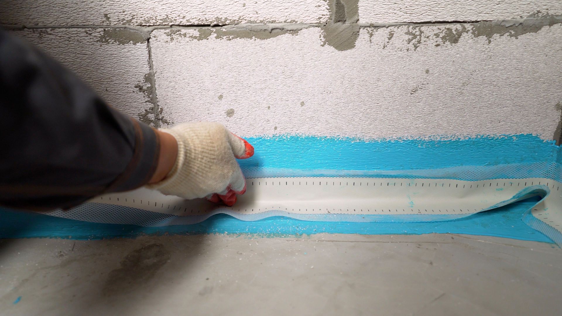 Person in gloves applying blue waterproofing tape to a concrete wall.