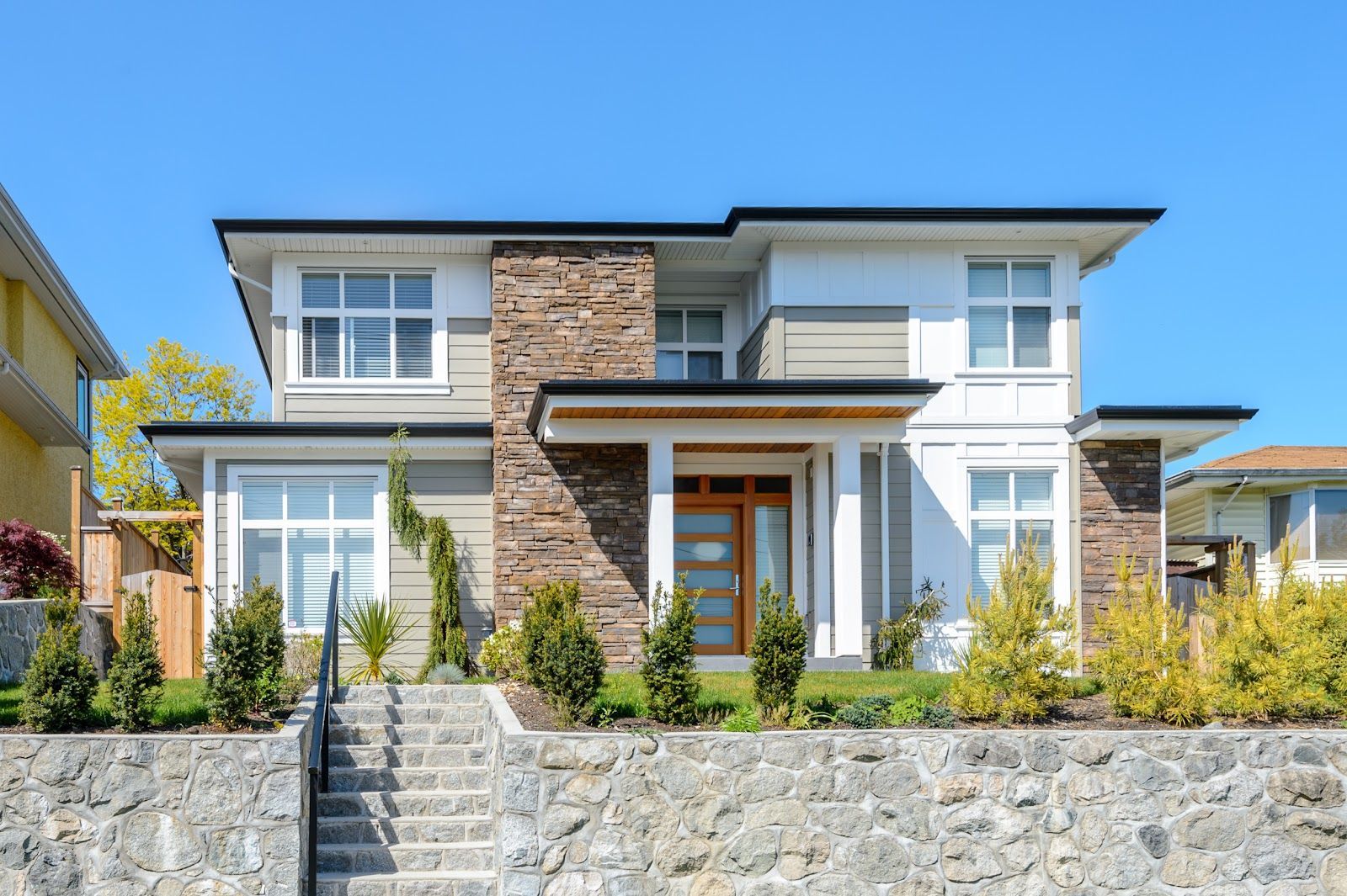 Modern two-story house with stone facade, light siding, and manicured landscaping under a blue sky.