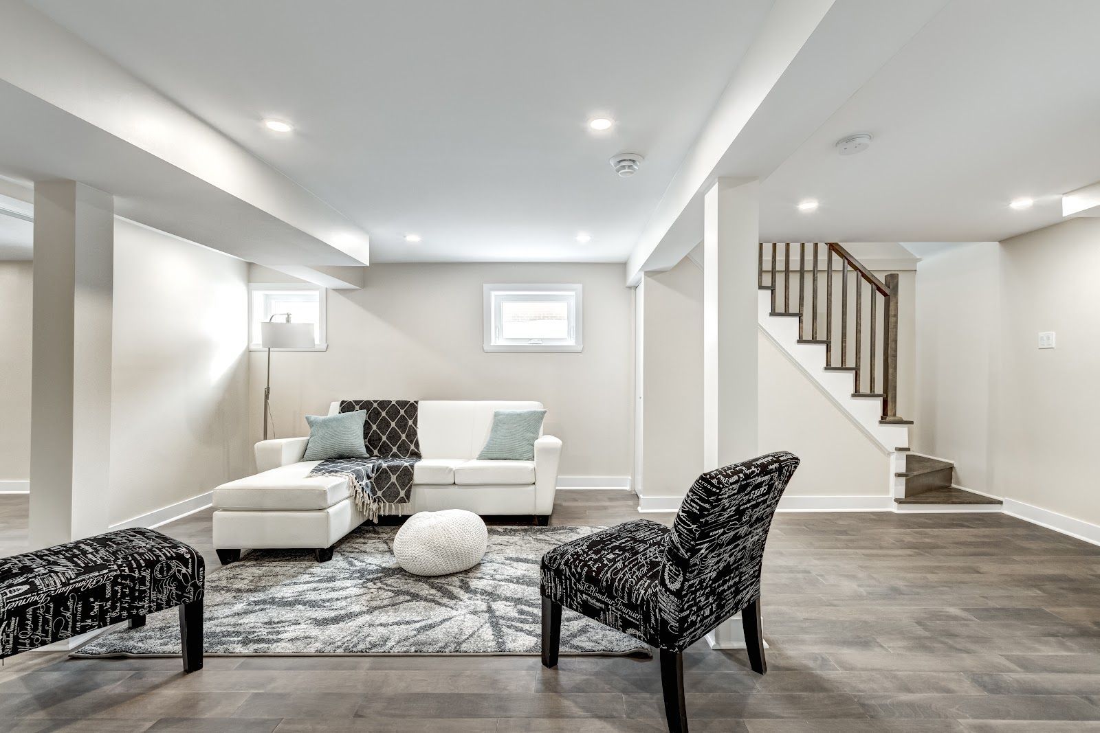 A finished basement with white walls, sectional sofa, patterned rug, and patterned armchairs. A staircase is visible.