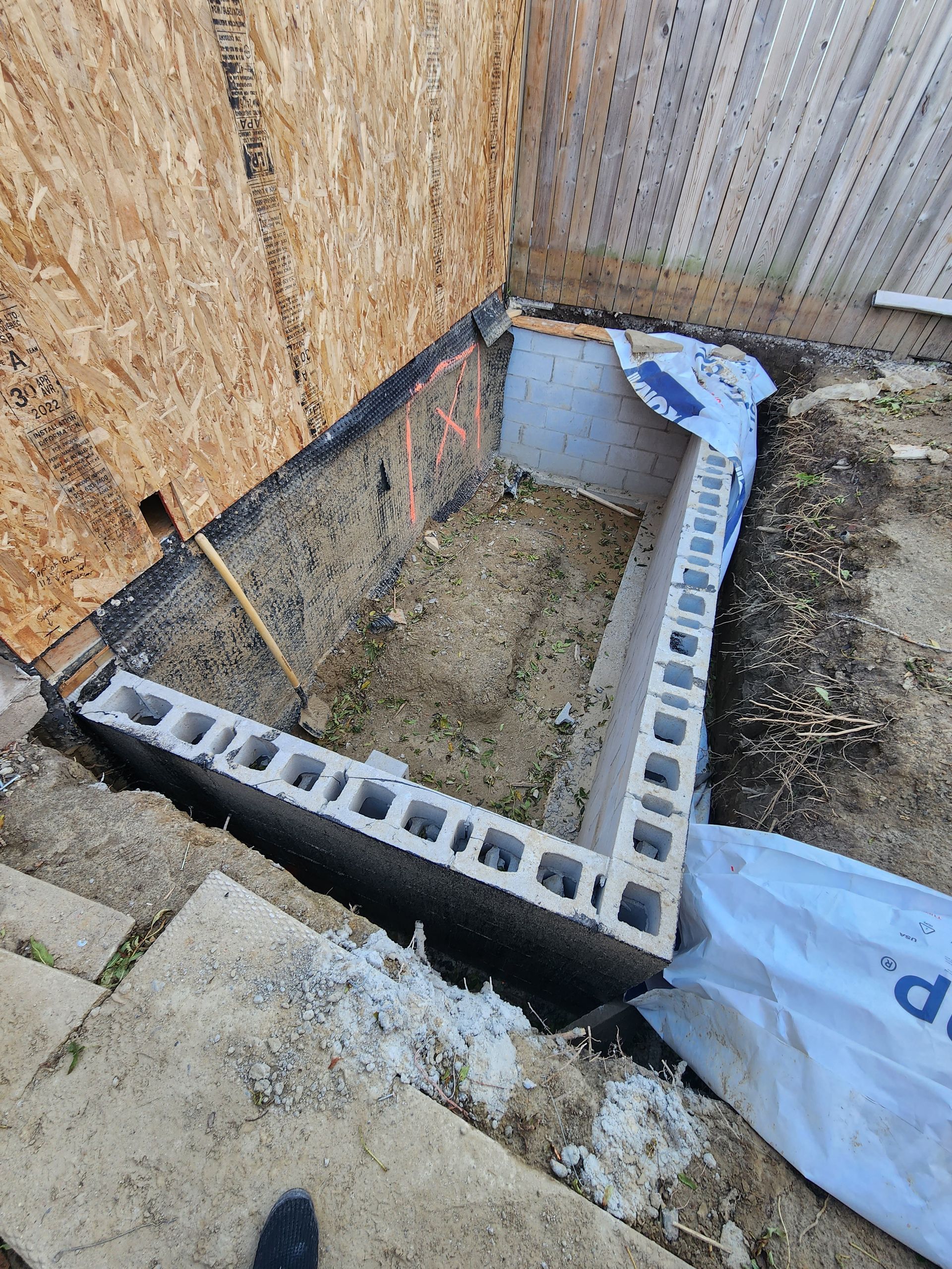 Construction site: concrete block walls forming a rectangular structure in a dirt yard, with a wooden fence in the background.