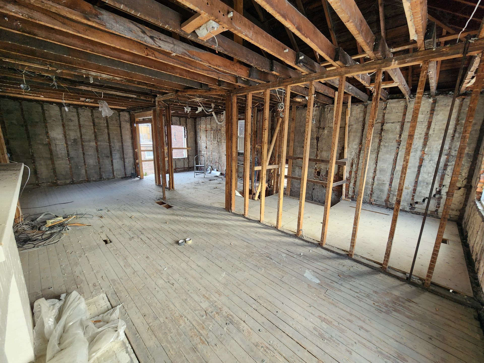 Interior view of a gutted room with exposed studs and beams. Debris covers the wooden floor.