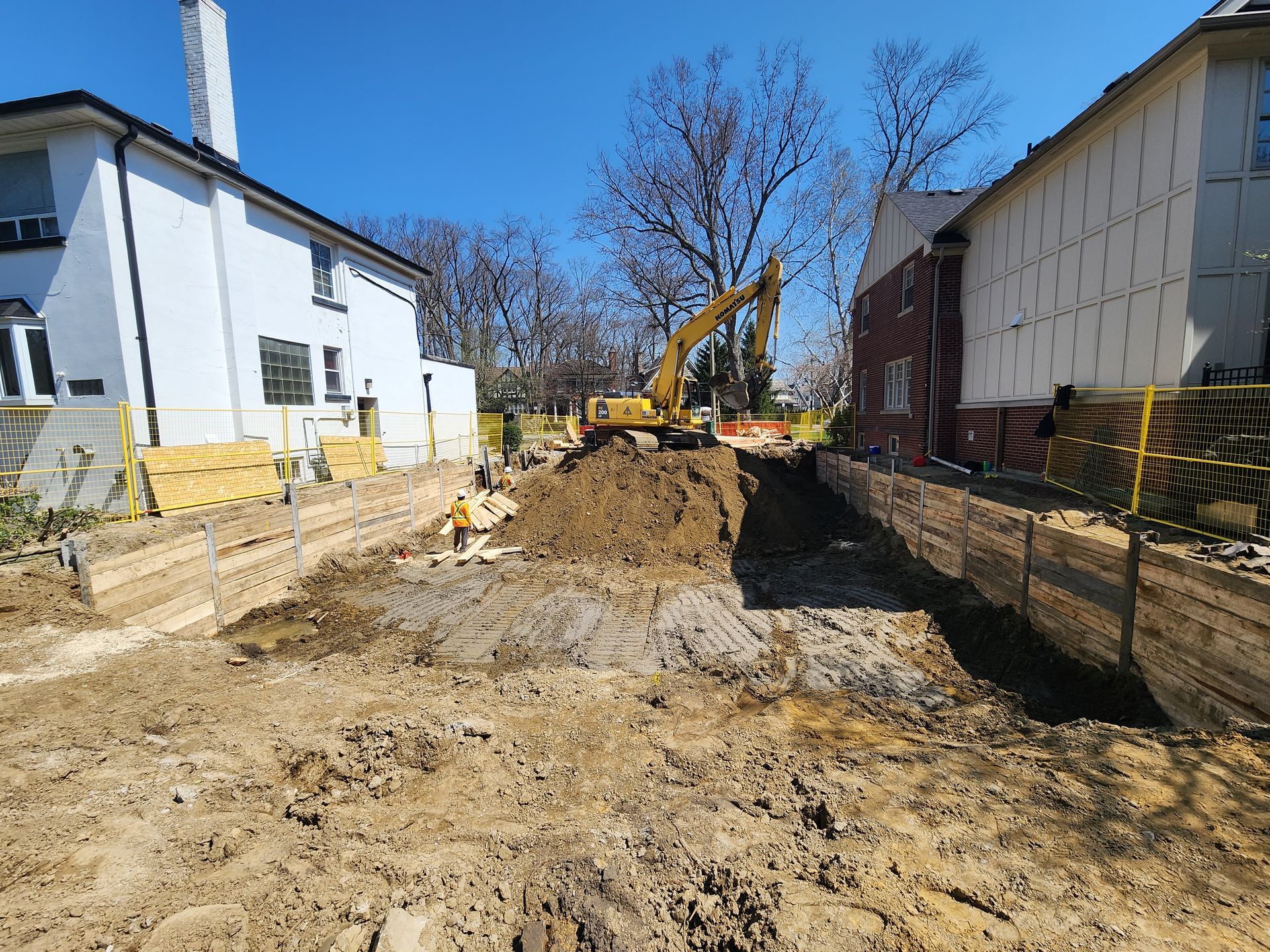 Construction site with excavator digging in a dirt pit between two houses. Yellow barriers and retaining walls visible.