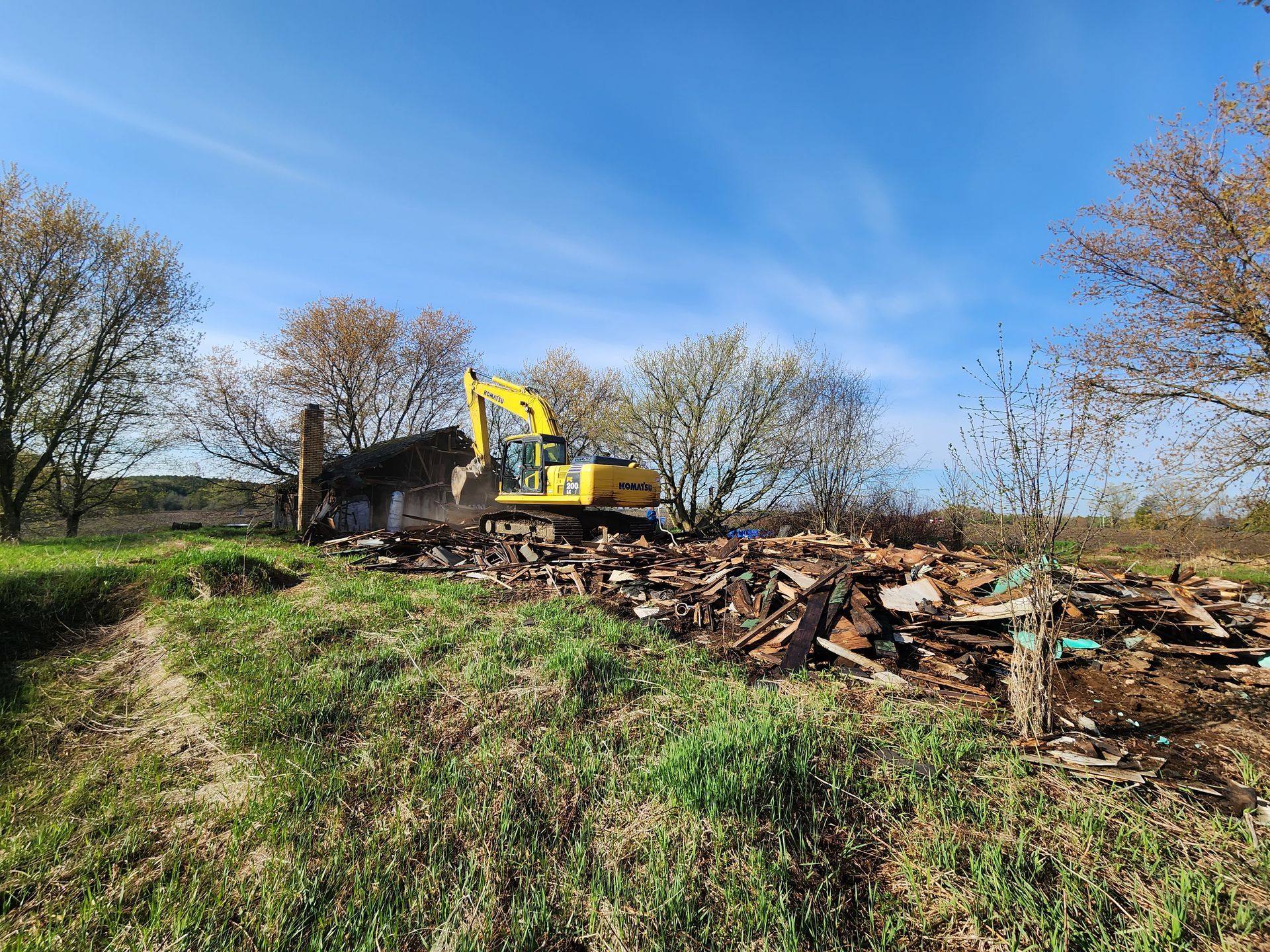 Yellow excavator demolishing a building on a grassy hill, blue sky background. Debris pile in the foreground.